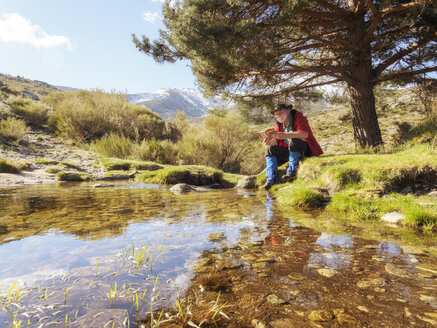 Spain, Sierra de Gredos, Hiker sitting at lake reading a book - LAF001653