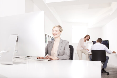 Geschäftsfrau arbeitet am Schreibtisch im Büro, während sich ihre Kollegen im Hintergrund unterhalten - MFRF000691