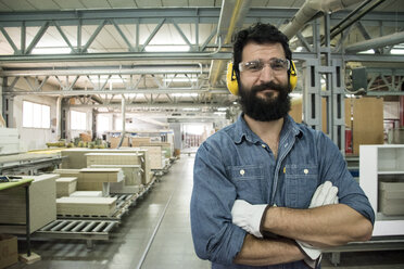 Mann mit Gehörschutz, Schutzbrille und Handschuhen lächelt mit verschränkten Armen in einer Fabrik - ABZF000602