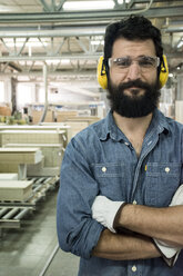 Mann mit Gehörschutz, Schutzbrille und Handschuhen lächelt mit verschränkten Armen in einer Fabrik - ABZF000603