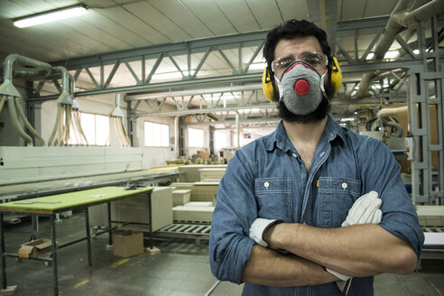 Mann mit Gehörschutz, Maske, Schutzbrille und Handschuhen mit verschränkten Armen in einer Fabrik - ABZF000605