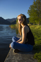 Italy, Lecco, teenage girl sitting on the edge of the lake at evening sunlight - MRAF000062