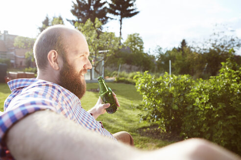 Junger Mann sitzt auf einer Bank im Garten und trinkt ein Bier - SEGF000565