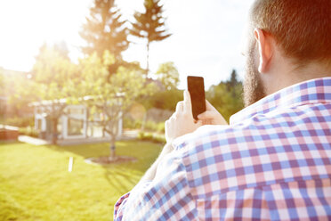 Junger Mann mit Smartphone im Garten, der seinen Garten fotografiert - SEGF000566