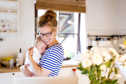 Glückliche Mutter mit Baby in der Küche und Blick auf den Laptop - HAPF000468