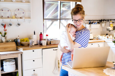 Glückliche Mutter mit Baby in der Küche und Blick auf den Laptop - HAPF000469