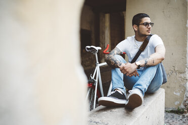 Junger Mann mit Fahrrad an der Wand sitzend - GIOF001163