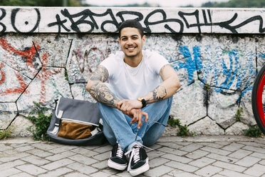 Smiling young man sitting at graffiti wall - GIOF001183