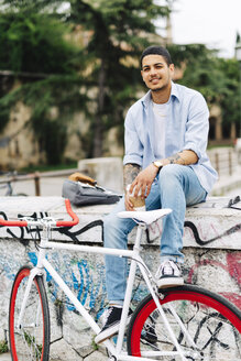 Lächelnder junger Mann mit einem Fahrrad auf einer Graffiti-Wand sitzend - GIOF001188