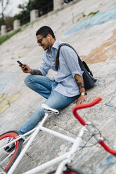 Junger Mann mit einem Fahrrad, der auf einer Mauer sitzt und auf sein Handy schaut - GIOF001201