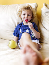 Porträt eines kleinen Jungen mit einem Lutscher und einem Apfel auf der Couch sitzend - MGOF001884