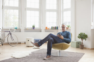 Älterer Mann sitzt zu Hause auf einem Stuhl und benutzt ein digitales Tablet - RBF004539