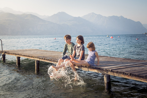 Italien, Brenzone, drei Kinder sitzen nebeneinander auf einem Steg und planschen mit Wasser, lizenzfreies Stockfoto