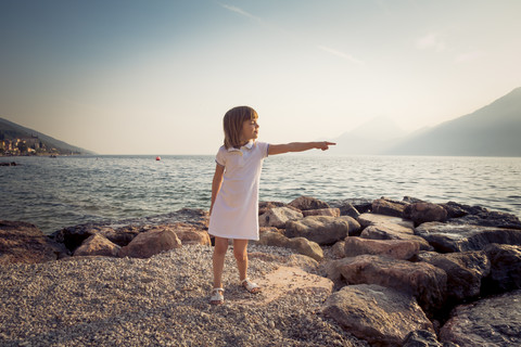 Italien, Brenzone, kleines Mädchen steht am Seeufer und zeigt auf etwas, lizenzfreies Stockfoto