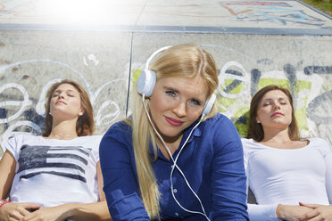 Lächelnde junge Frau, die mit Kopfhörern Musik hört, während sich ihre Freunde im Hintergrund entspannen - FMKF002705