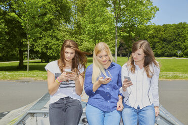 Three friends sitting side by side using their smartphones - FMKF002711