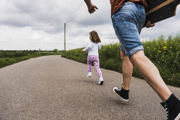 Father with skateboard and daughter running on country lane - UUF007405