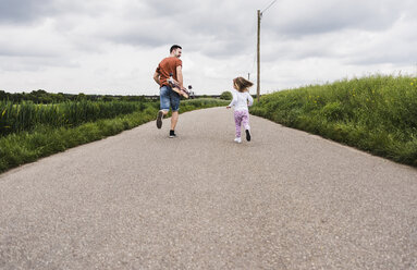 Father with skateboard and daughter running on country lane - UUF007406
