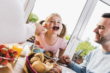 Mädchen mit Familie leckt Löffel am Frühstückstisch - UUF007423