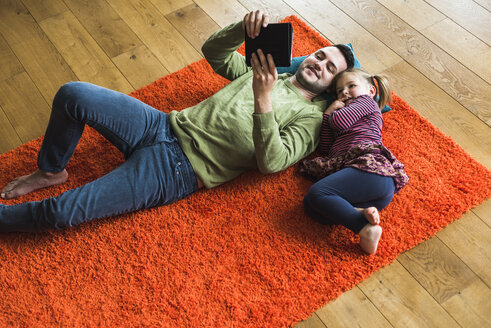 Vater und Tochter liegen auf einem Teppich auf dem Boden und benutzen ein digitales Tablet - UUF007466