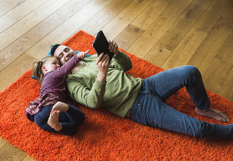 Vater und Tochter liegen auf einem Teppich auf dem Boden und benutzen ein digitales Tablet - UUF007467