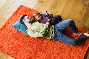 Vater und Tochter liegen auf einem Teppich auf dem Boden und benutzen ein digitales Tablet - UUF007471