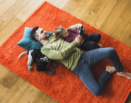 Vater und Tochter liegen auf einem Teppich auf dem Boden und benutzen ein digitales Tablet - UUF007472