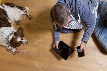 Draufsicht auf einen Mann, der auf dem Boden neben einem Hund sein Smartphone benutzt - MAEF011756