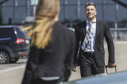 Lächelnder Geschäftsmann mit Gepäck auf einem Parkplatz, der eine Frau ansieht - MADF000946