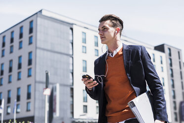 Young man with cell phone, earbuds and folder on the move - UUF007526