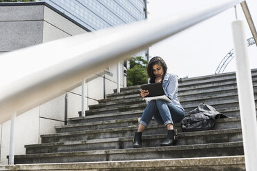 Junge Frau sitzt auf einer Treppe und schaut auf ein digitales Tablet - UUF007533