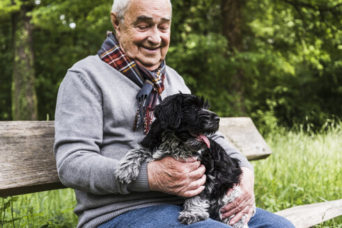 Lächelnder älterer Mann sitzt mit seinem Hund auf einer Bank in der Natur - UUF007567