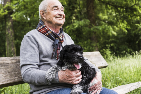 Lächelnder älterer Mann sitzt mit seinem Hund auf einer Bank in der Natur - UUF007568