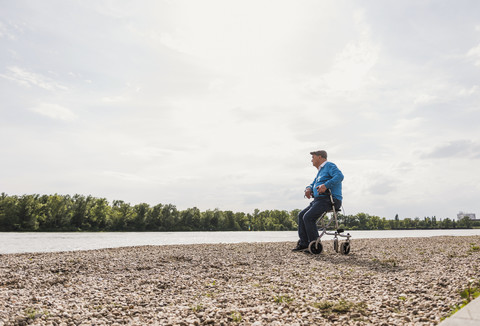 Älterer Mann sitzt auf einem Rollator am Flussufer, lizenzfreies Stockfoto