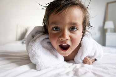 Porträt eines kleinen Jungen mit Duschtuch auf dem Bett liegend, der nach einem Bad lustige Gesichter zieht - VABF000517