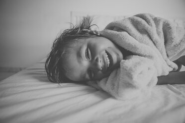 Porträt eines lachenden kleinen Jungen mit Duschtuch auf dem Bett liegend nach einem Bad - VABF000518