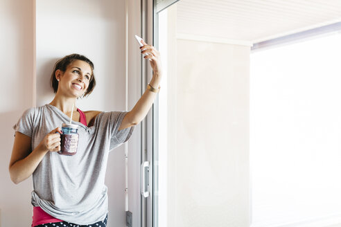 Lächelnde junge Frau mit gesundem Getränk macht ein Selfie am Fenster - JRFF000738