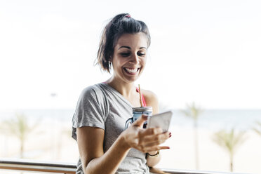 Glückliche junge Frau mit gesundem Getränk und Smartphone auf dem Balkon - JRFF000744