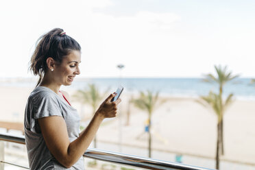 Lächelnde junge Frau schaut auf dem Balkon auf ihr Smartphone - JRFF000745