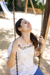 Laughing young woman sitting on a swing at playground - VABF000569