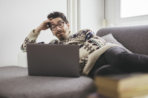 Verwunderter Mann liegt auf einer Couch und schaut auf einen Laptop, lizenzfreies Stockfoto