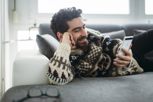 Smiling man lying on a couch looking at phablet - LCUF000022