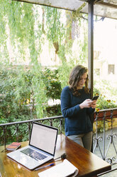 Young woman sitting on balcony using laptop and smart phone - BOYF000376