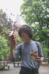 Deutschland, Berlin, junger Mann mit Brezel schaut auf sein Smartphone - RTBF000225