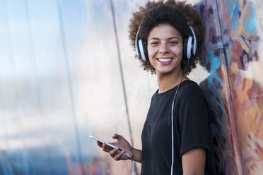 Portrait of happy young woman with headphones and smartphone leaning against wall - SIPF000538