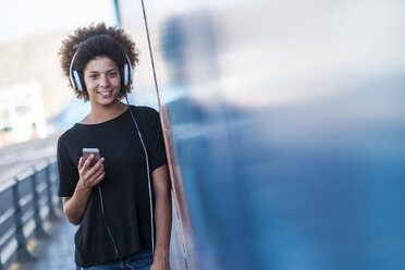 Portrait of smiling young woman with headphones and smartphone leaning against wall - SIPF000539