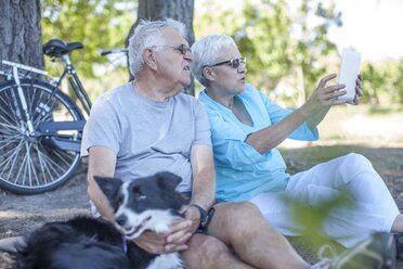 Elderly couple sitting outdoors with dog and digital tablet - ZEF008706