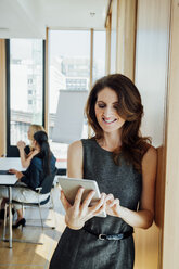 Smiling businesswoman in office using digital tablet with meeting in background - CHAF001747