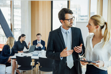 Businessman and businesswoman talking in office with meeting in background - CHAF001751