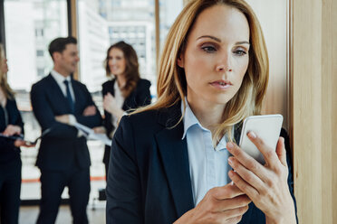 Geschäftsfrau im Büro schaut auf ein Handy mit Geschäftsleuten im Hintergrund - CHAF001784